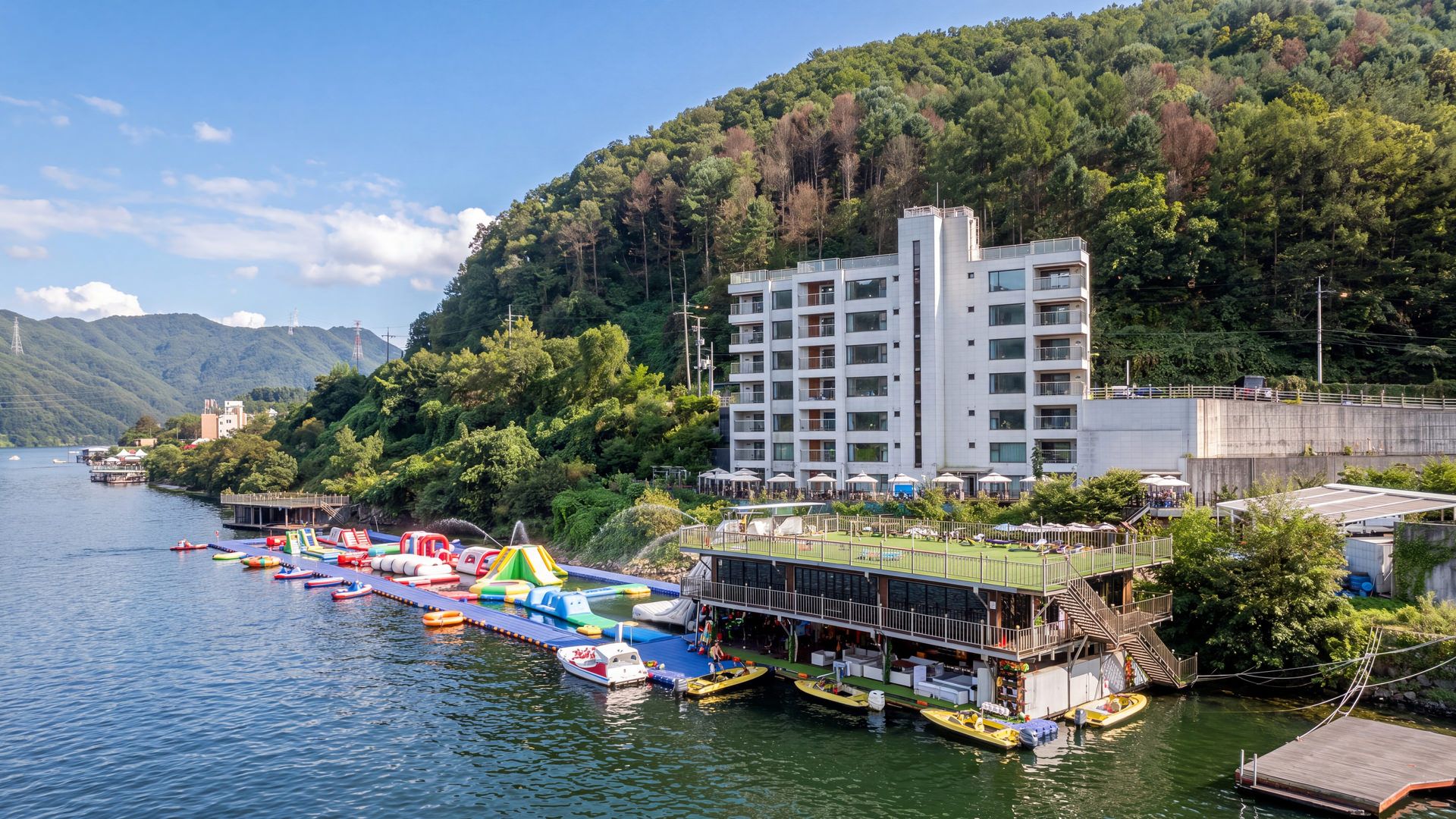 Un hotel frente al agua con zona de piscina, barcos y una ladera cubierta de bosque.