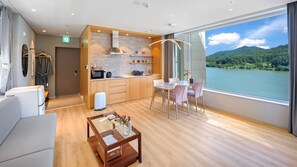 A modern hotel room with a wooden floor, a dining area with a table and chairs, a kitchenette, a large window with a view of a lake and mountains, and a bathroom with a shower and toilet. - Gapyeong Laputa Resort (Gapyeong)