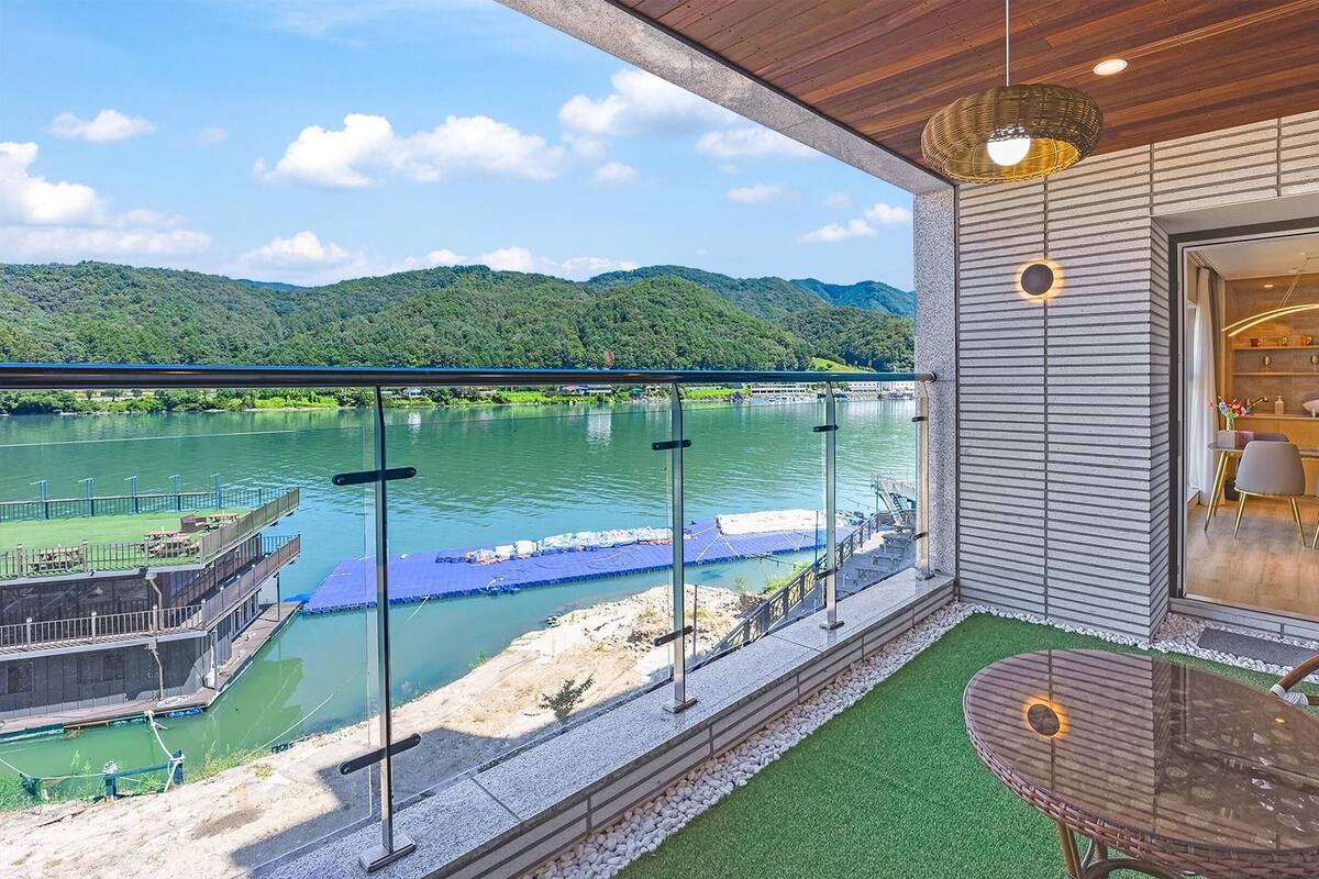 Ein Balkon mit Glasgeländer, der auf einen Fluss blickt, mit Tisch und Stühlen, sowie einer Aussicht auf eine Brücke und Hügel.