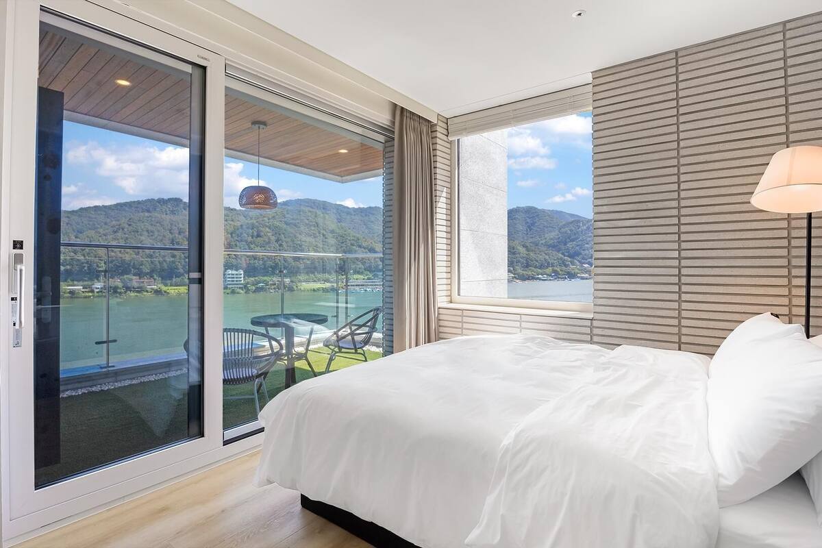 Ein Schlafzimmer mit Bett, Nachttisch, Lampe, einem Fenster mit Blick auf einen See und Berge sowie einer Glastür, die auf einen Balkon mit Tisch und Stühlen führt.