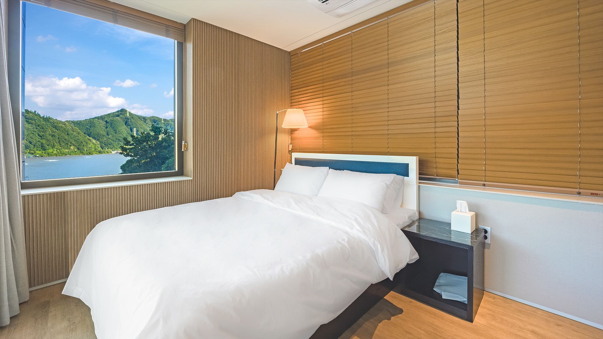 Habitación de hotel con cama, mesita de noche con lámpara, ventana con vistas a un lago y colinas, y cabecero de madera.