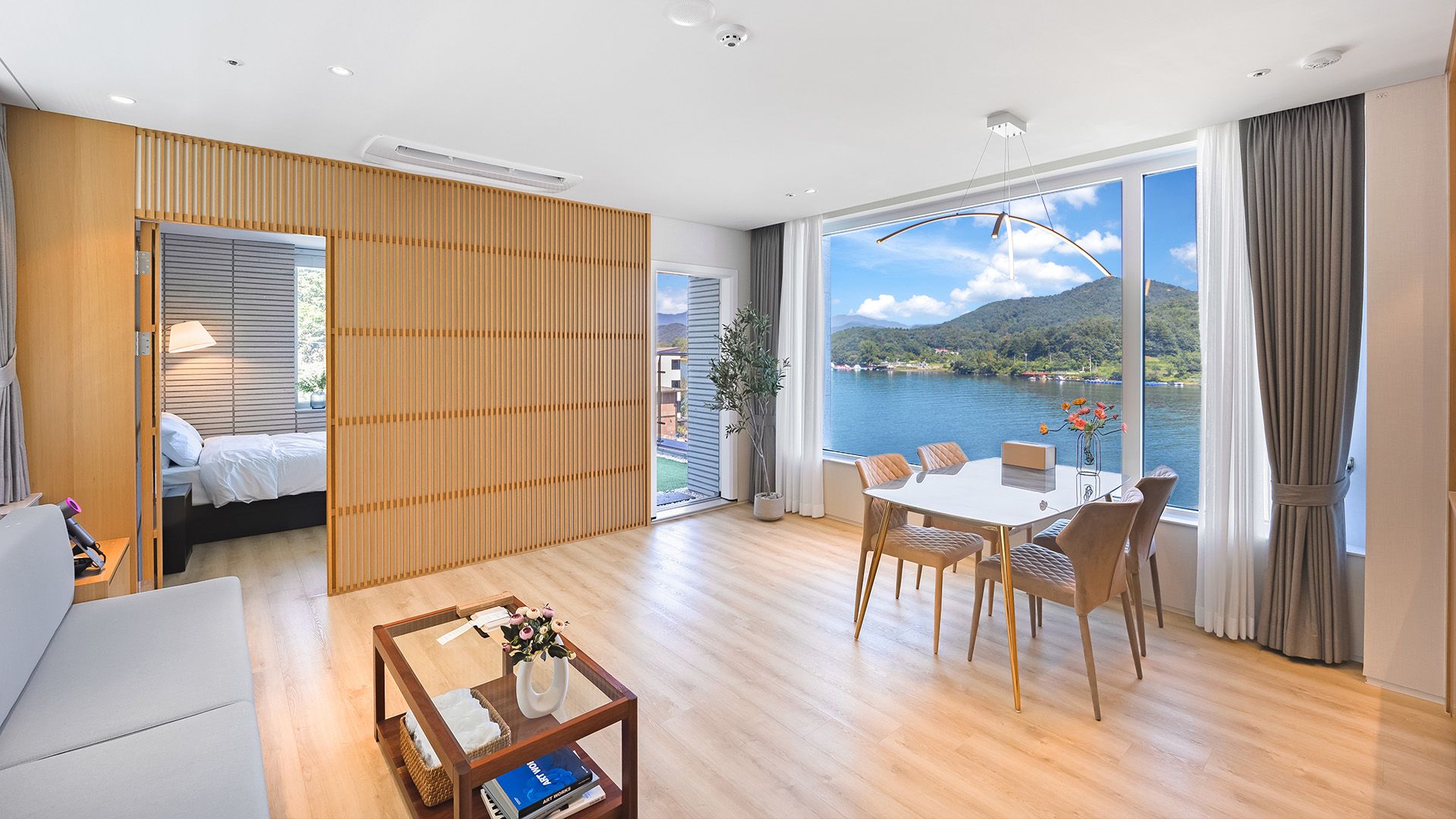 A modern hotel room with a bed, a dining area, and a large window offering a lake view.
