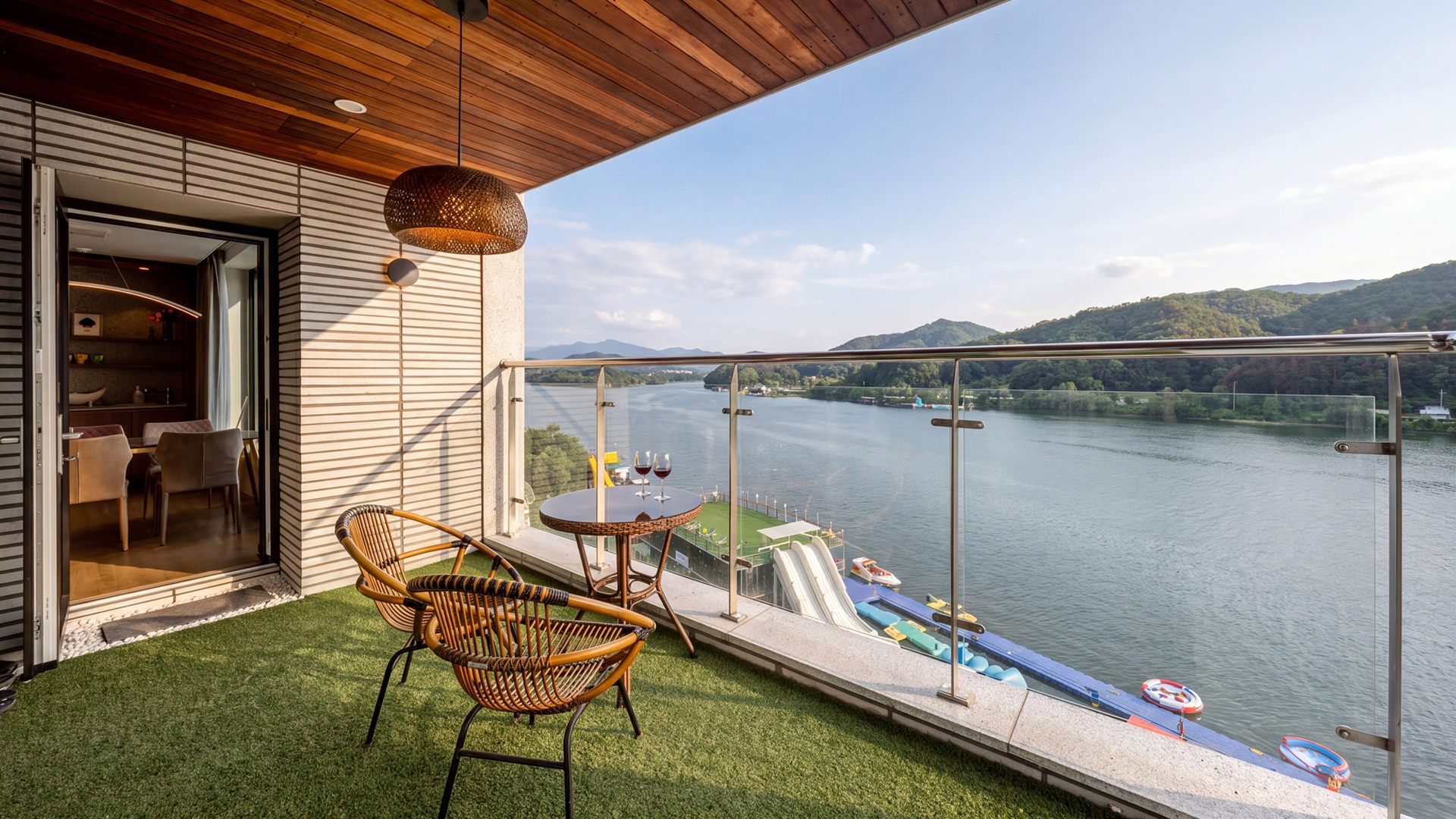A balcony with a view of a river and mountains, furnished with wicker chairs and a table, and a glass railing.