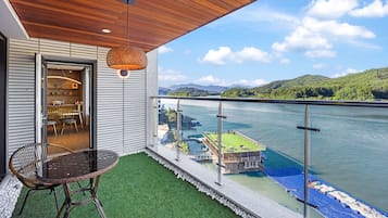 A balcony with a view of a river and mountains, a wicker chair, and a table.