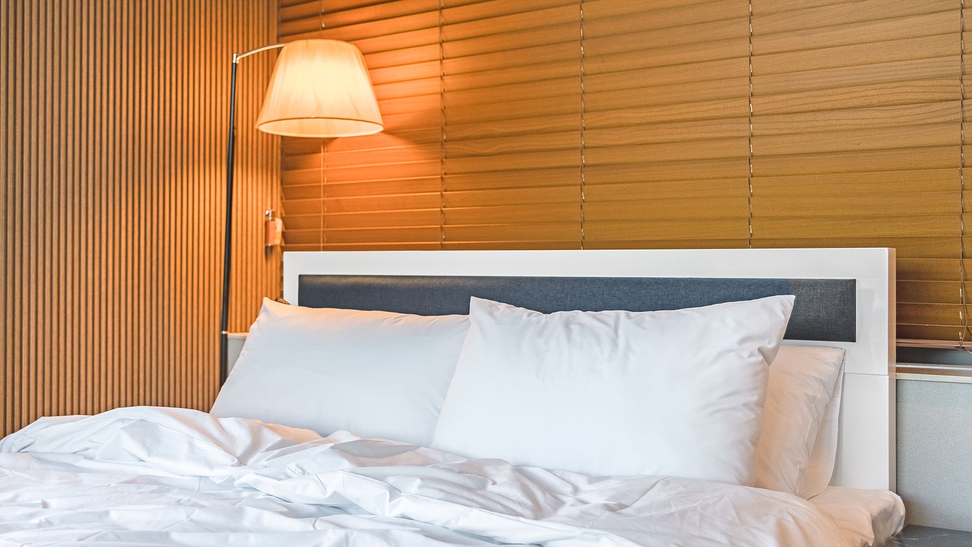 A neatly made bed with white linens and pillows, a bedside lamp, and wooden slatted blinds.
