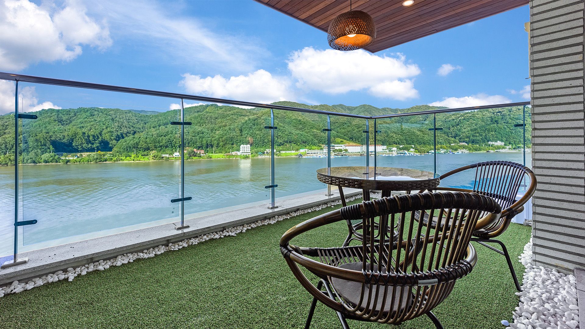 A balcony with a glass railing, a table, and two wicker chairs overlooking a lake with a forested hillside in the background.
