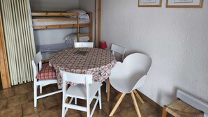 Huoneessa on kerrossänky, ruutukuviollinen pöytäliina peitetty pöytä, kaksi valkoista tuolia ja kaksi kehystettyä kuvaa seinällä.