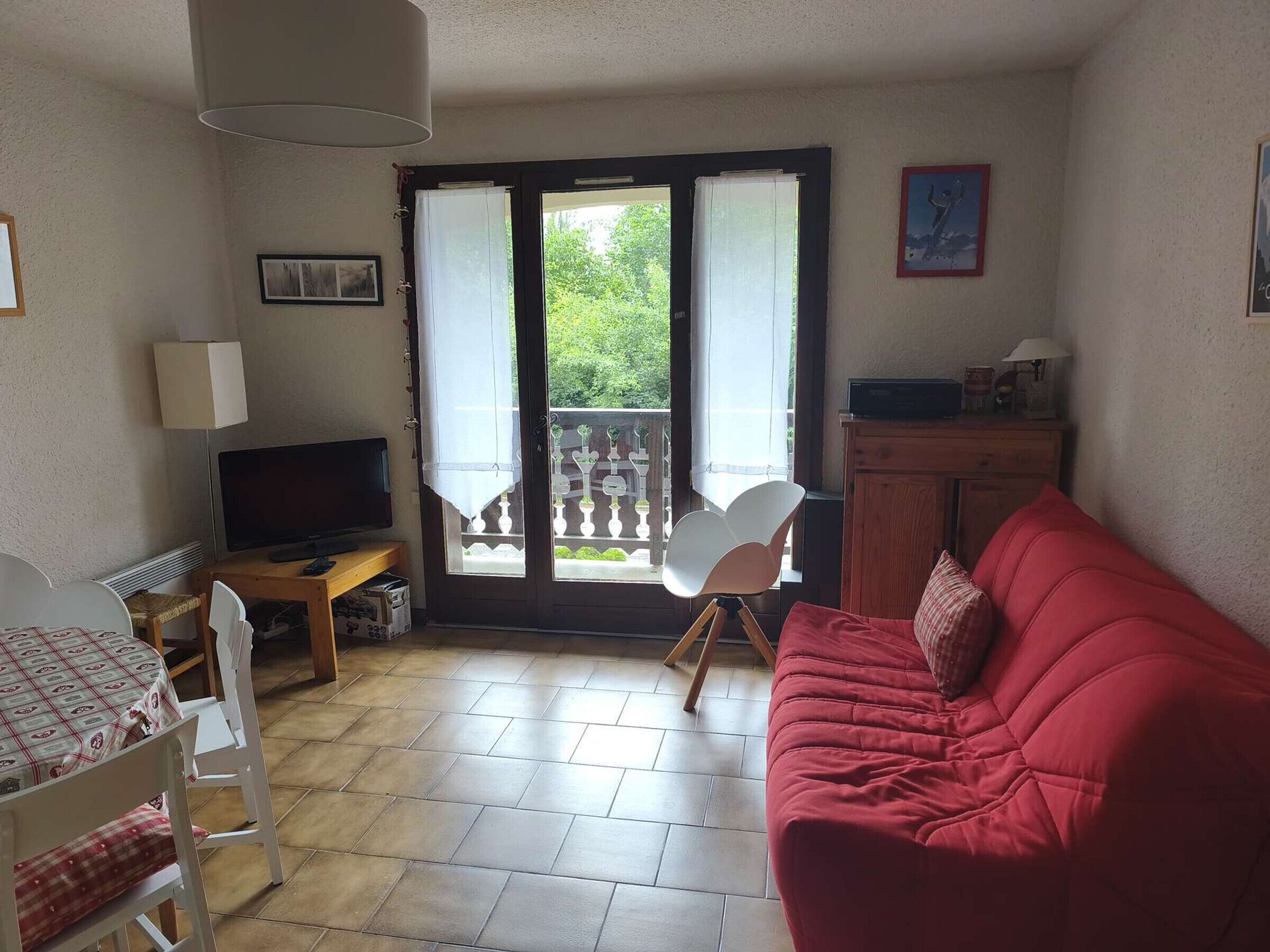 A living room with a red sofa, a TV, a dining table with chairs, a balcony with a chair, and a cabinet.