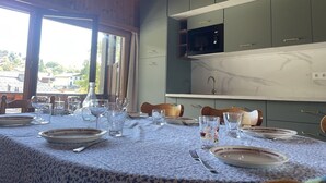 Un comedor con una mesa preparada para cuatro, una cocina con armarios verdes y un microondas, y vistas a casas a través de amplios ventanales.