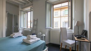 Hotellihuone, jossa on sänky, työpöytä tietokoneella, tuoli, patteri ja ikkuna, josta on näkymä rakennuksiin.