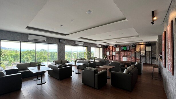 A spacious lounge area with multiple seating options, large windows offering a view of greenery, and a modern design with a mix of wood and metal elements.