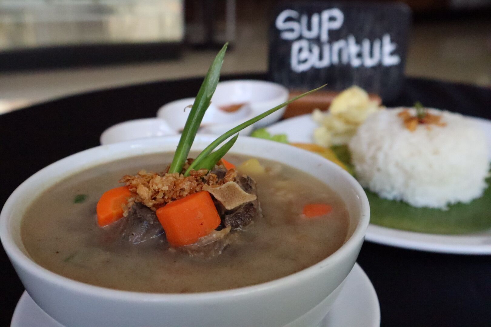 A bowl of soup with meat and vegetables, a plate of rice, and a sign that reads "Sup Burit".
