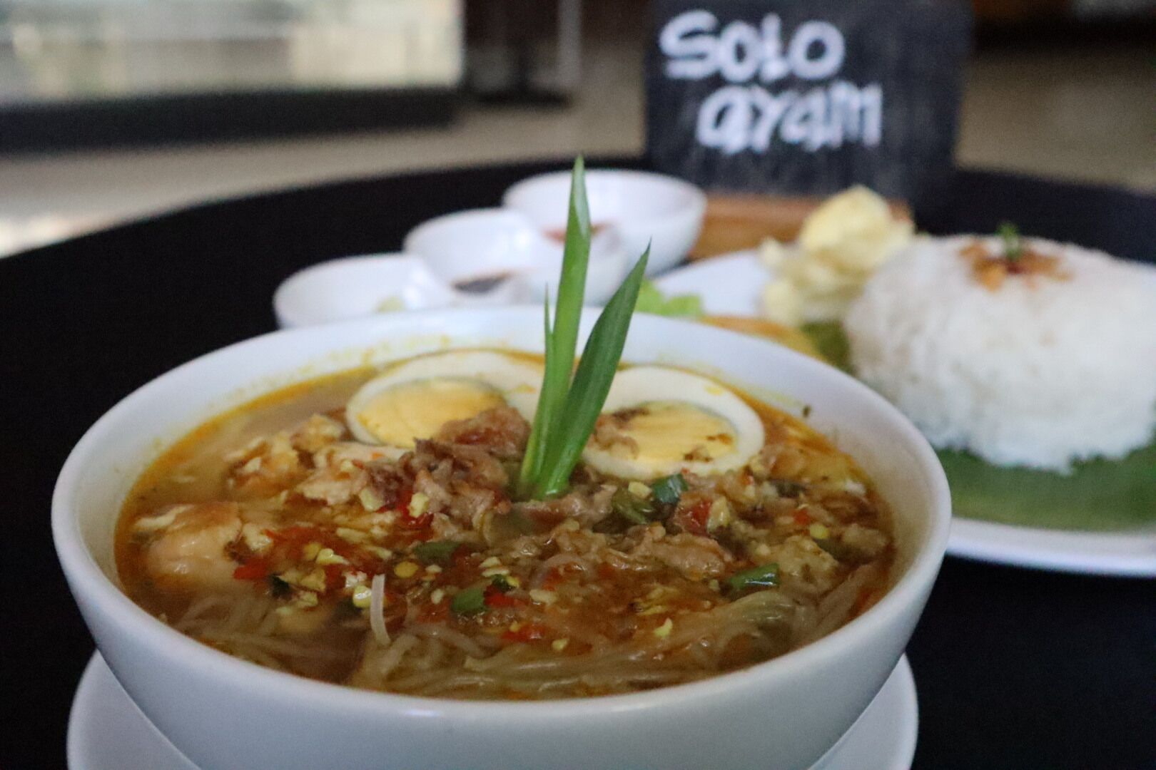 A bowl of soup with meat and vegetables, a boiled egg, and a side of rice on a plate.