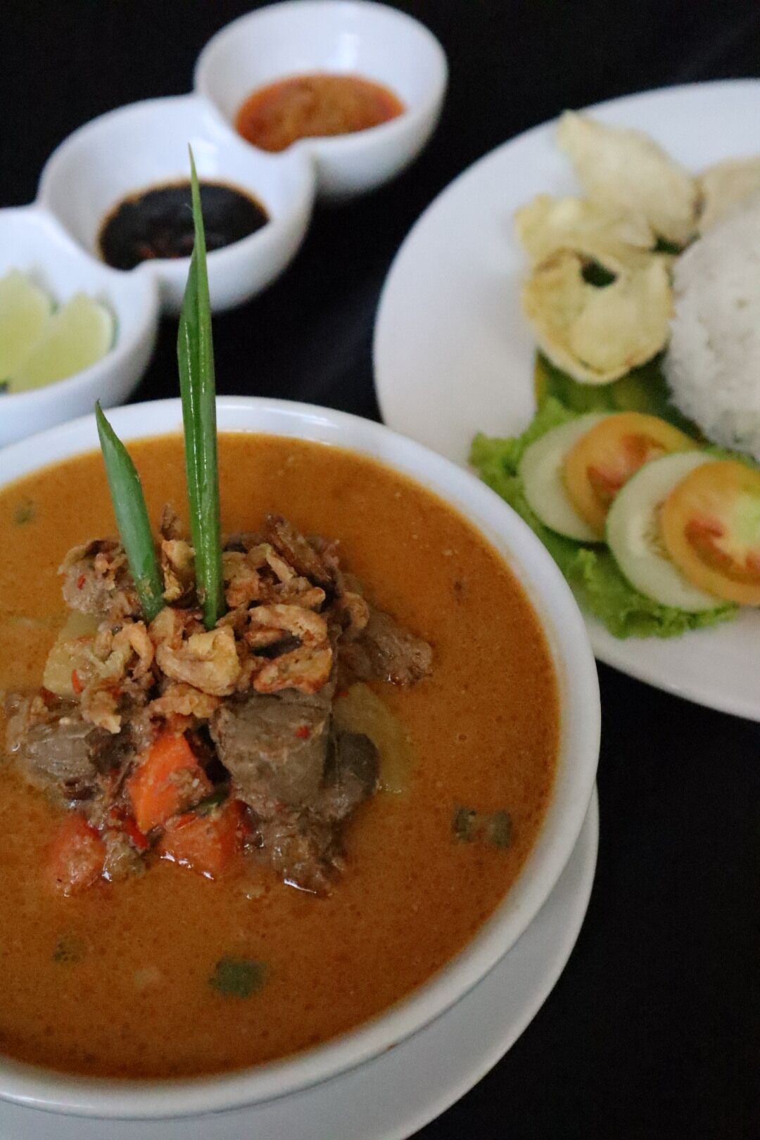 A bowl of curry with meat and vegetables, accompanied by rice and a salad with sliced vegetables.