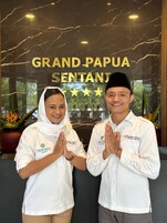 Dos personas con camisas blancas y el logotipo de Grand Papua Sentai, de pie frente a una pared de mármol con el nombre del hotel.