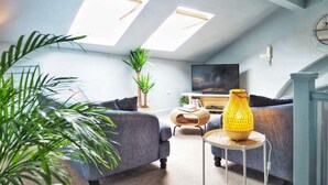 Ein modernes Wohnzimmer mit hohen Decken, zwei Oberlichtern, einem grauen Sofa, einem kleinen runden Tisch mit einer gelben Lampe und einem Fernseher auf einem Schrank.
