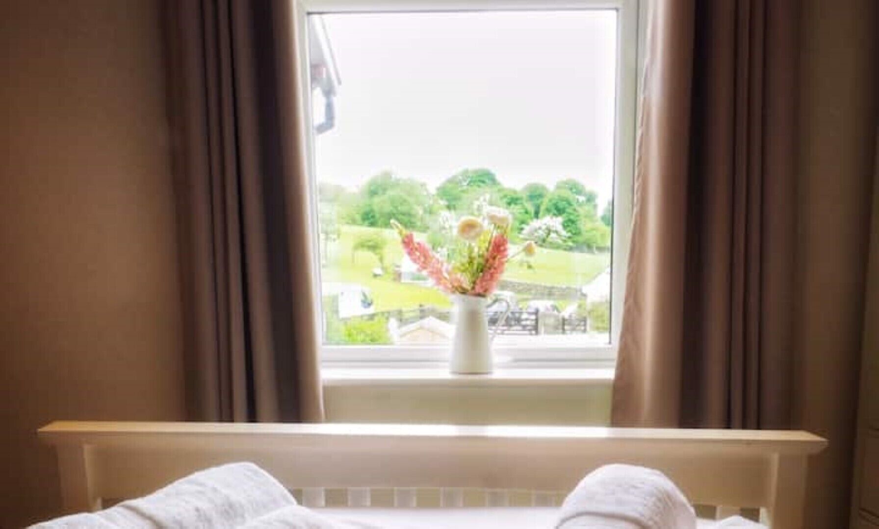 Une pièce avec un lit, une fenêtre avec des rideaux et un vase de fleurs posé sur le rebord de la fenêtre.