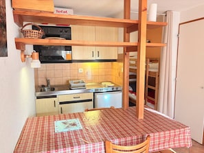 A compact kitchen with a microwave, stove, and sink, a checkered table, and a wooden shelf with a basket.