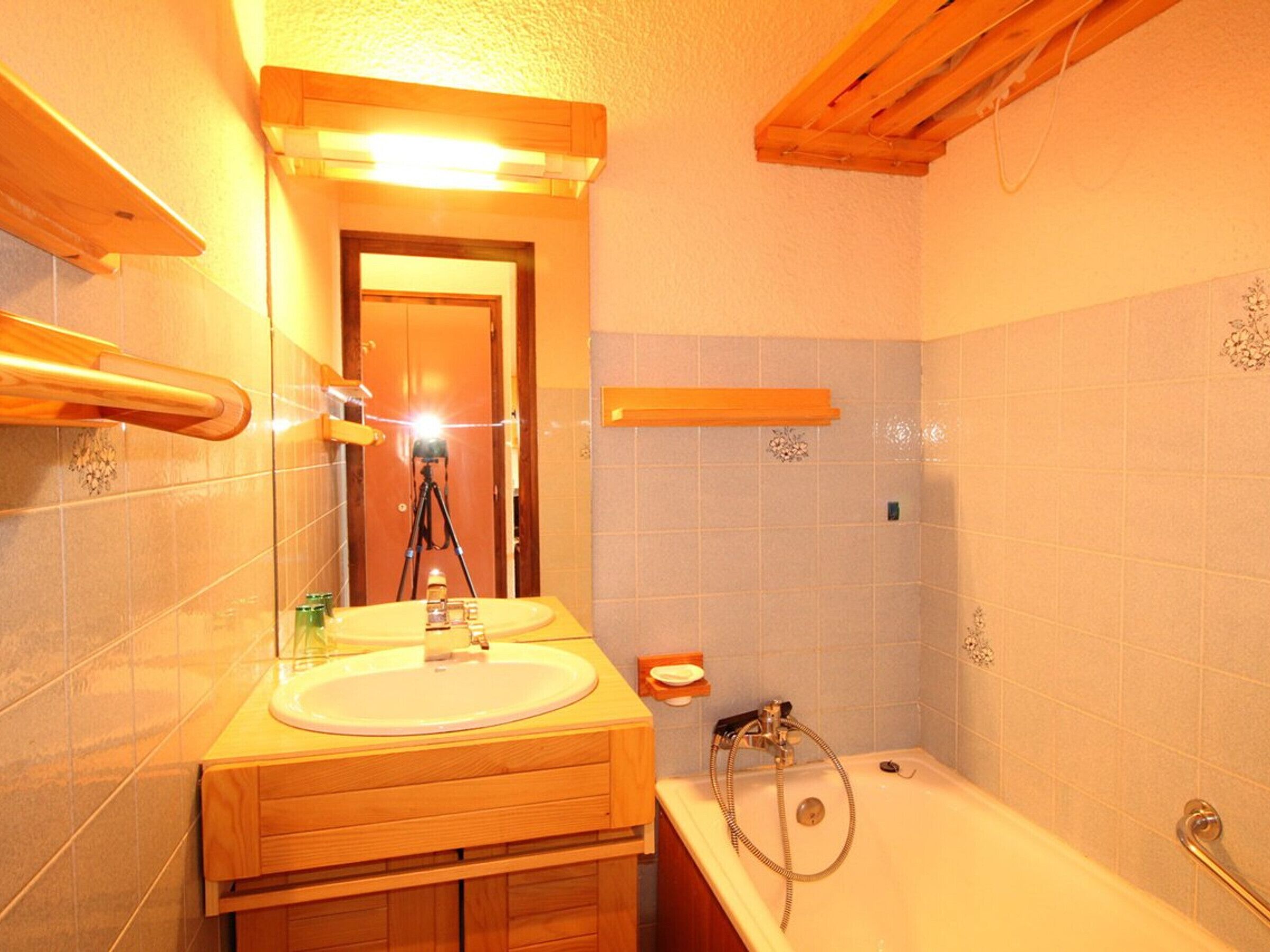 Une salle de bain avec un meuble-lavabo en bois, un évier, un miroir, une douche équipée d’un pommeau de douche à main et une porte visible au fond.