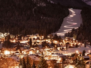 Une ville de montagne enneigée, illuminée la nuit, avec des bâtiments éclairés et une route sinueuse.
