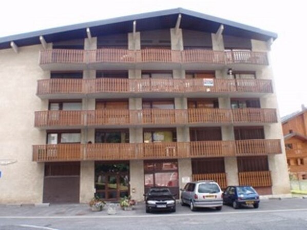 A multi-story building with balconies, a central entrance, and parked cars in front.