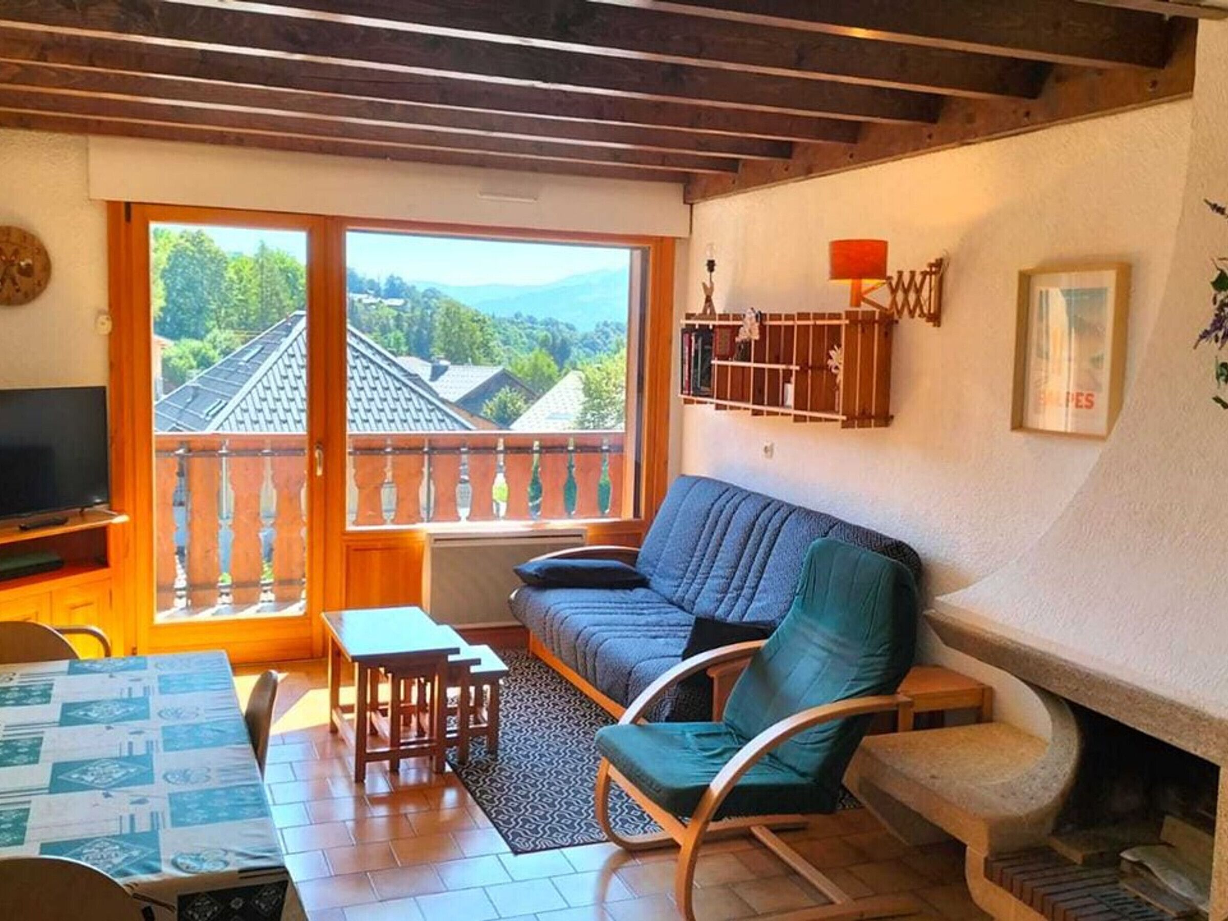 A cozy living room with a sofa, chairs, a small table, and a TV. There's a balcony with a view of houses and trees.