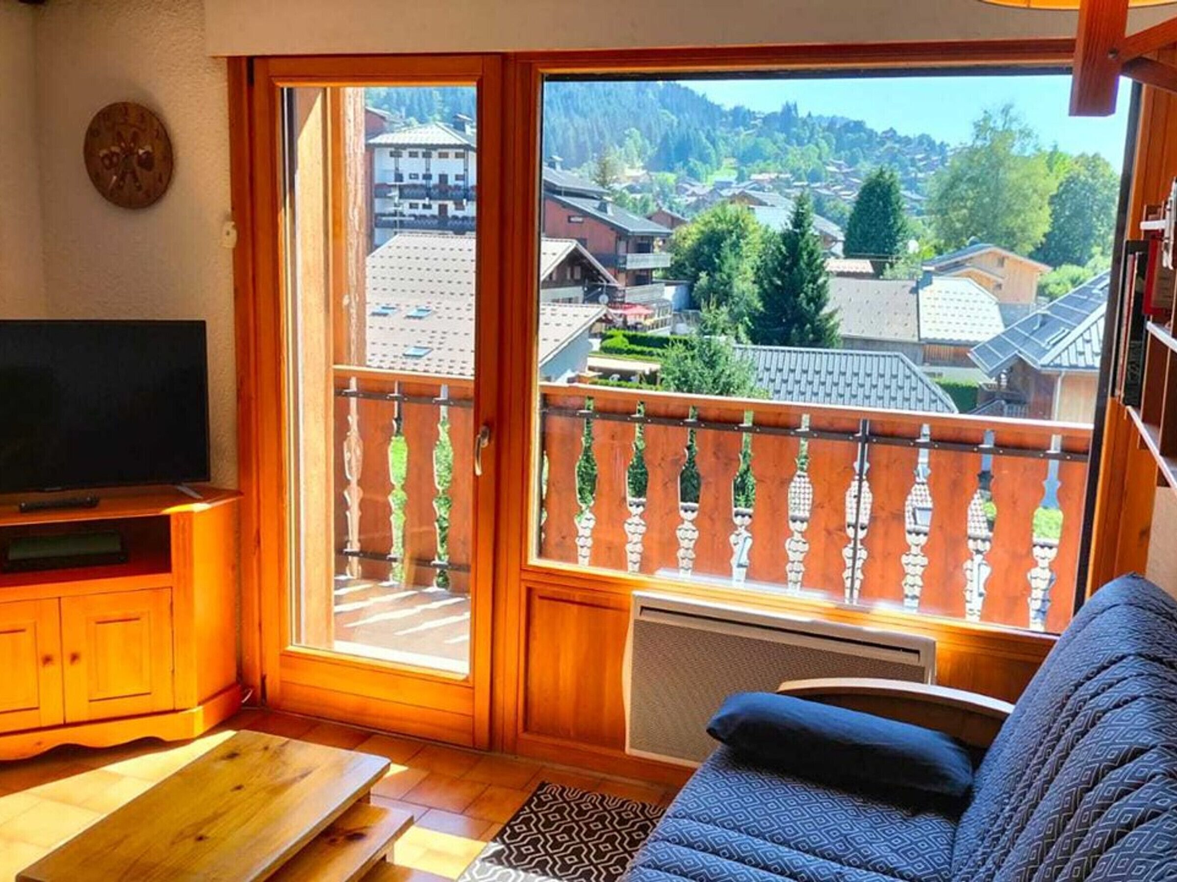 A room with a television, a sofa, a wooden table, and a large window with a view of a village and mountains.