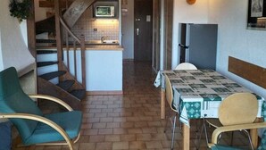 Kompakti keittiö, jossa on puinen portaikko, ruokailutila pöydän ja tuolien kera sekä pieni oleskelualue, jossa on yksi tuoli.