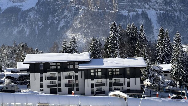 Ein Gebäude mit flachem Dach und zahlreichen Fenstern, umgeben von schneebedeckten Bäumen und Bergen.