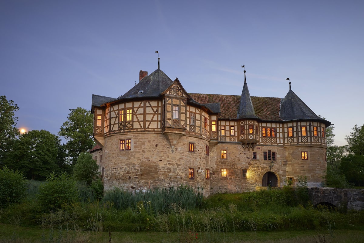A historic stone building with multiple towers and a turret, surrounded by lush greenery.