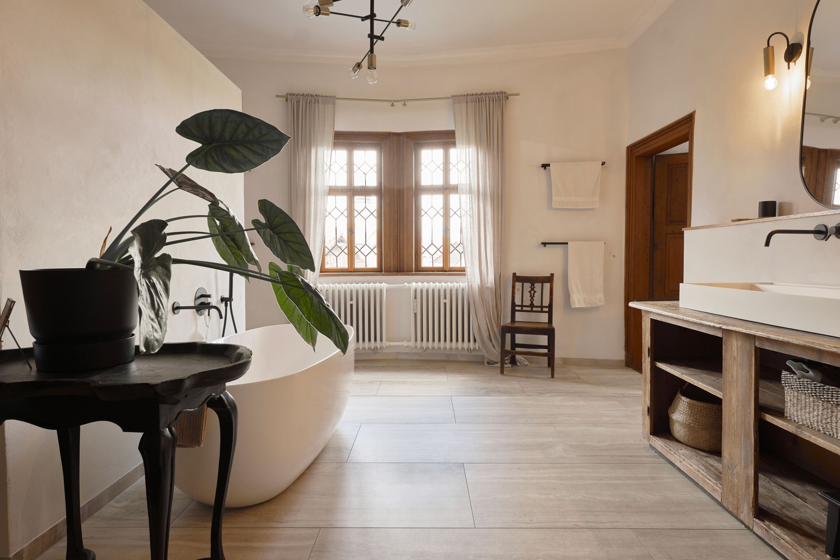 A bathroom with a bathtub, a wooden chair, a potted plant, a radiator, and a window with curtains.