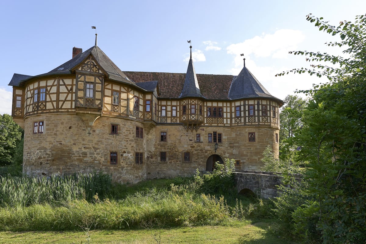 A large, historic stone building with multiple towers and a turret, surrounded by lush greenery.