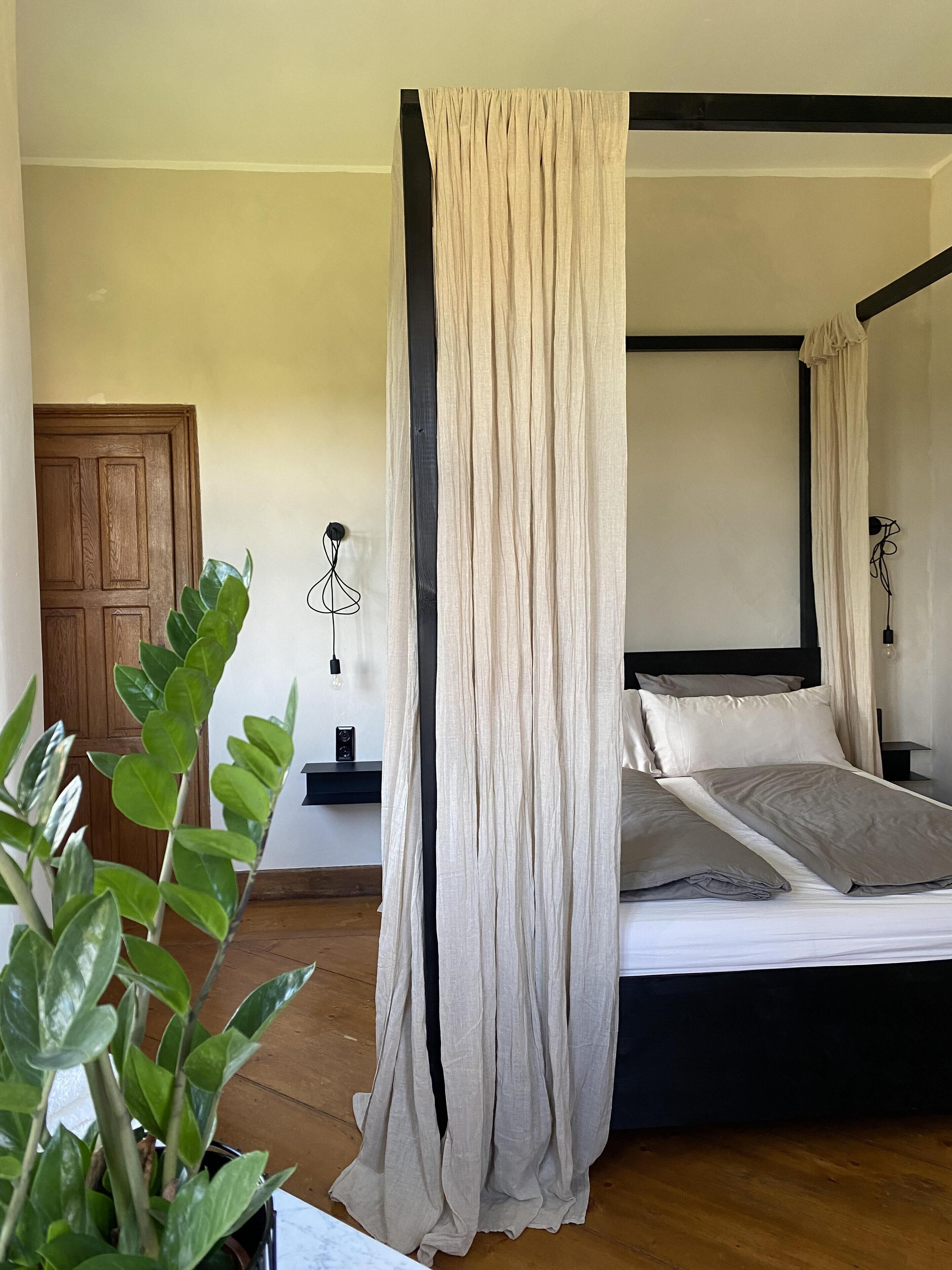 A bedroom with a four-poster bed, a wooden door, a bedside table with a lamp, and a potted plant.