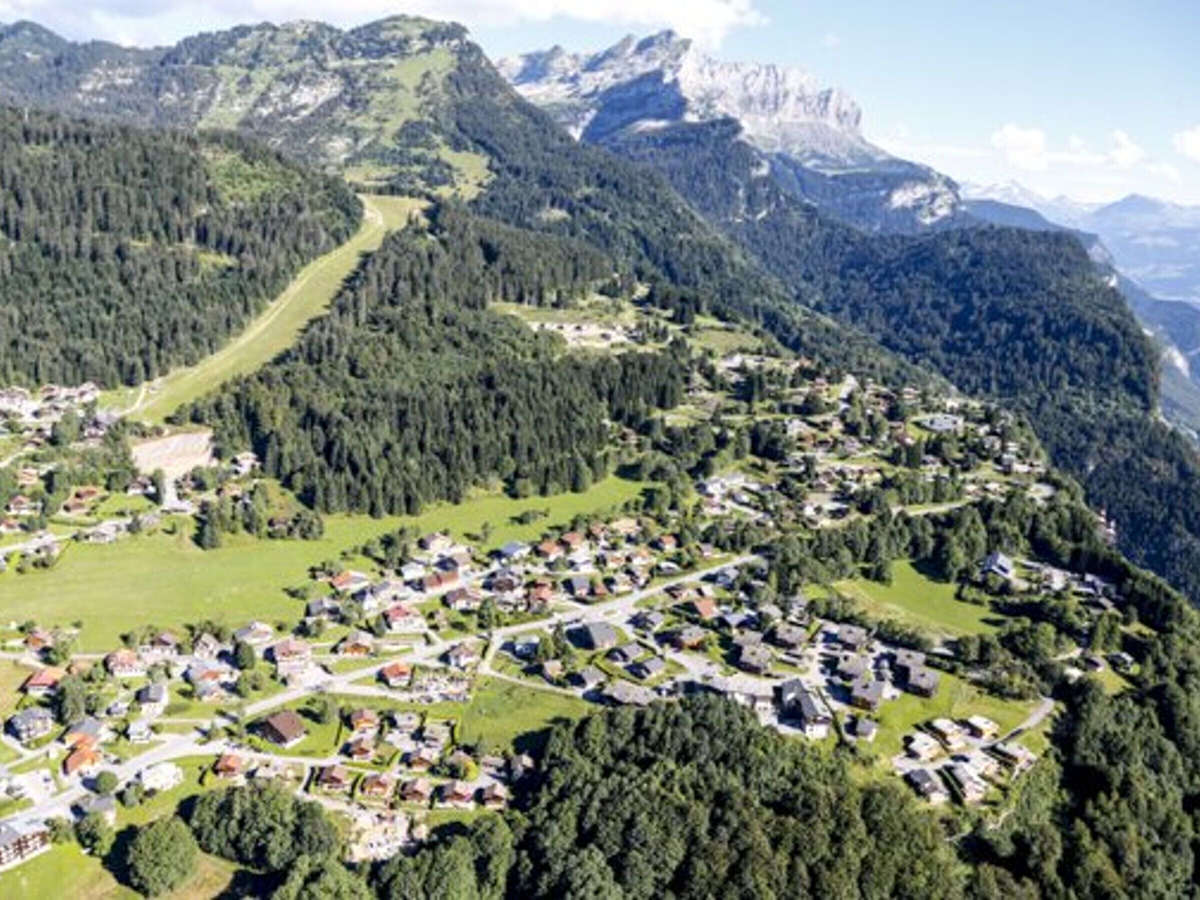 An aerial view of a mountain town with dense forested areas, a winding road, and scattered buildings.