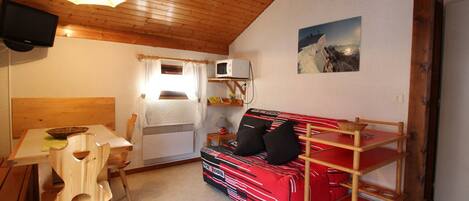 Ein gemütliches Zimmer mit Holzboden, einem roten Sofa auf Rollen, einem kleinen Tisch mit einer Schüssel, einem Sessel, einer Mikrowelle und einem Bild an der Wand.