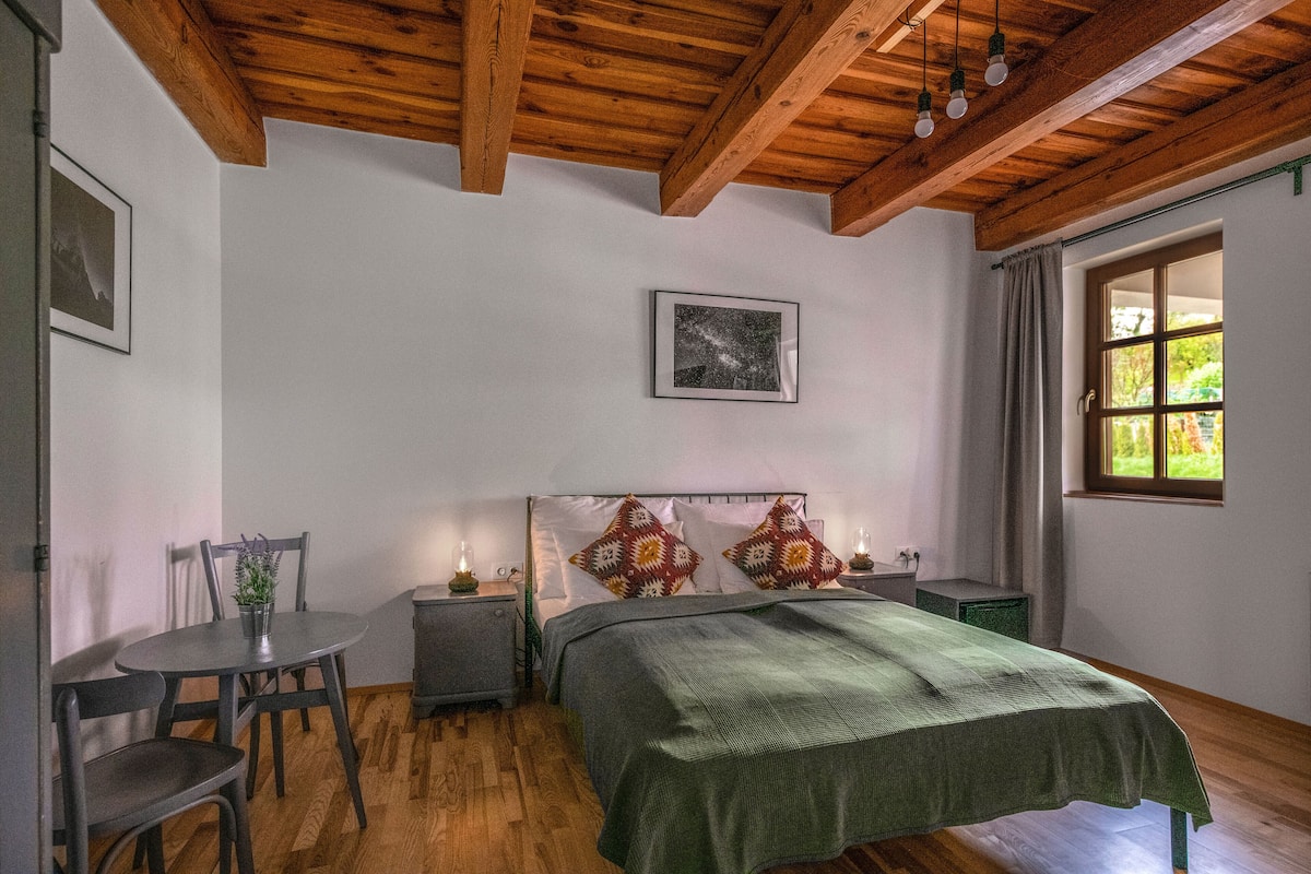 Un dormitorio con techo de madera, una cama con manta verde, una mesita de noche, una silla, una pequeña mesa con una planta y una ventana con cortinas.