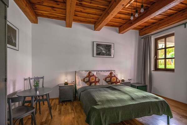 A bedroom with a wooden ceiling, a bed with a green blanket, a bedside table, a nightstand, a chair, a small table with a plant, and a window with curtains. - Ferrata Guesthouse (Csesznek)