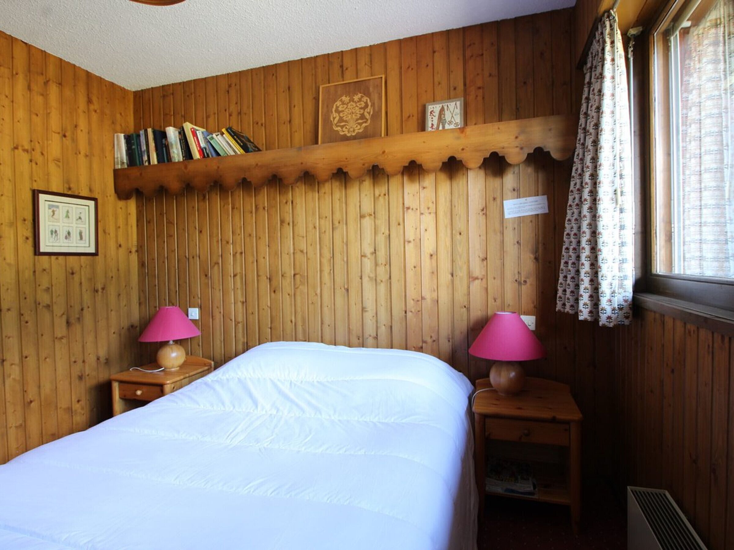 Ett rum med träpanel på väggarna, en säng, två sängbord med lampor, en hylla med böcker och ett fönster med gardiner.