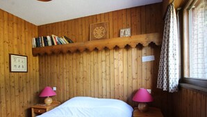 Une pièce aux murs lambrissés, comprenant un lit, deux tables de chevet avec des lampes, une étagère remplie de livres et une fenêtre avec des rideaux.