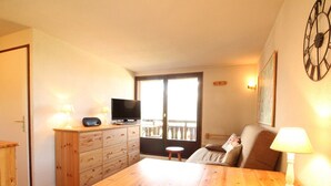 Una habitación acogedora con una mesa de madera, un televisor en un mueble, un balcón con barandilla y una lámpara sobre la mesa.