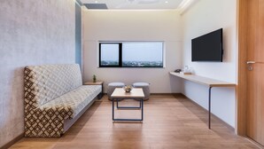 A modern living room with a sofa, a small table, two ottomans, a TV mounted on the wall, and a large window providing a view. - 7 Apple Hotel Nashik (Nashik)