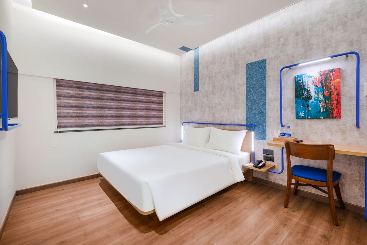 A modern hotel room with a bed, a desk with a chair, a window with blinds, and a ceiling fan.
