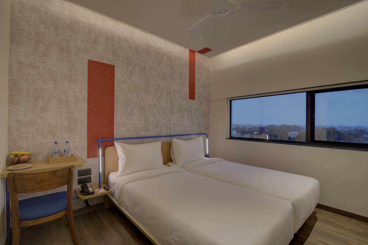 A modern hotel room with a single bed, a desk with a chair, a small table with a bowl of fruit, and a window with a city view.