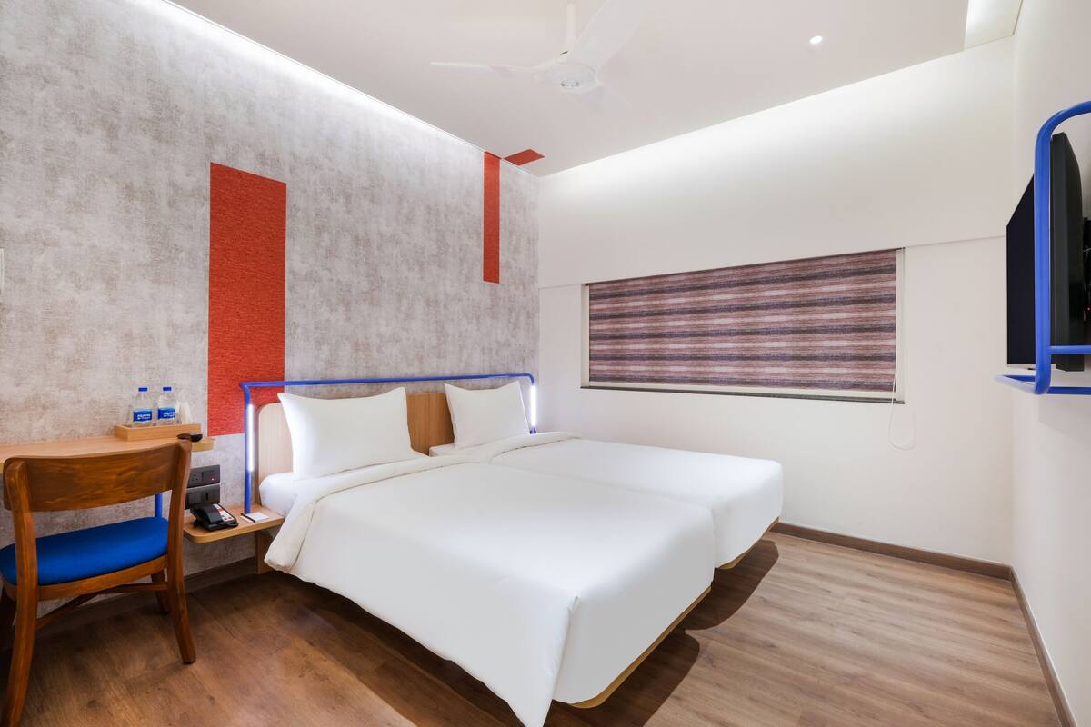A hotel room with a bed, a desk with a chair, a TV, and a window with blinds.