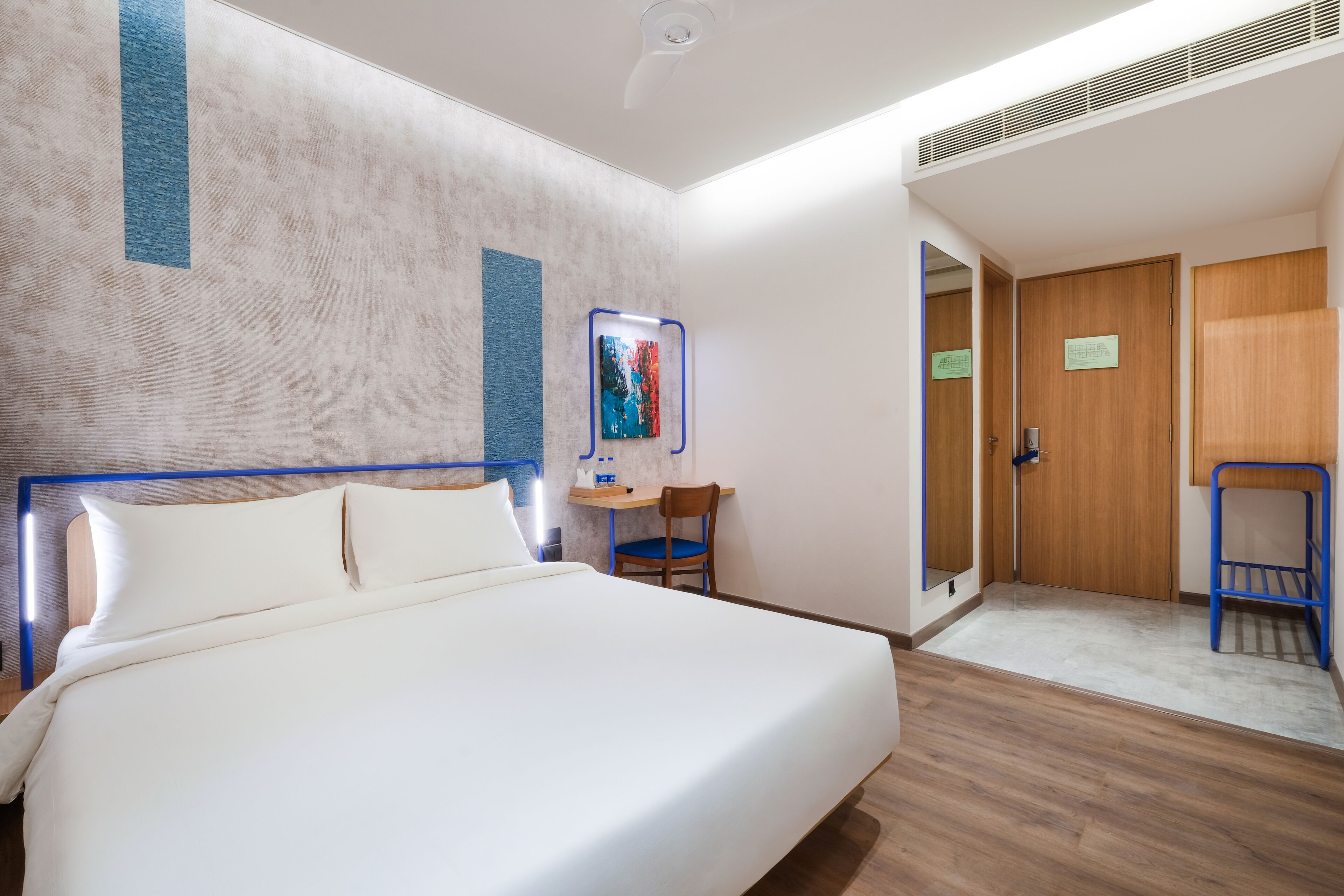 Quarto de hotel com uma cama grande, uma escrivaninha com cadeira, área de chuveiro e portas de madeira.