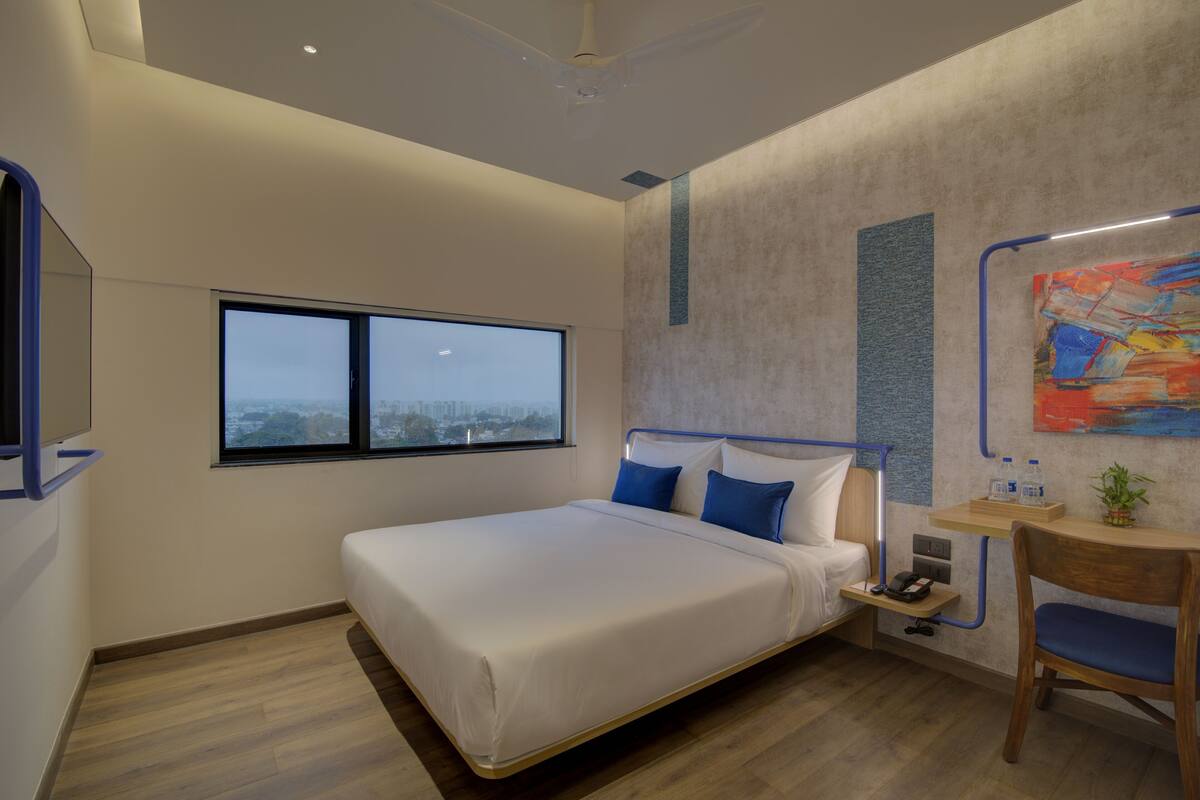 A modern hotel room with a large bed, a desk with a chair, a window, and a painting on the wall.