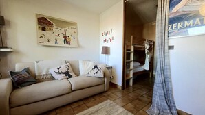 Un salón acogedor con un sofá, un tapete, un cuadro y una cama litera al fondo.