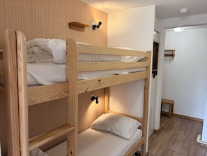A bunk bed with a wooden frame and white bedding, a small shelf, and a wall-mounted light fixture.