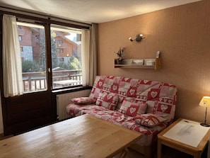A cozy room with a wooden table, a red and white patterned sofa, a small side table with a lamp, and a window with curtains.