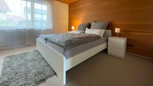 A bedroom with a wooden ceiling, a bed with a grey and white duvet, a bedside table with a lamp, and a window with a view of a car outside.